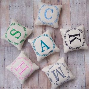 May include: A collection of small, square decorative pillows. Each pillow features a different embroidered letter within a wreath of leaves. The letters are in various colors, including blue, green, pink, and black, on a cream-colored background.