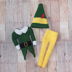 May include: A felt elf costume set, including a green jacket with white collar and cuffs, yellow pants, and a green hat with a yellow band and red feather. The costume is designed for a doll or small figure.