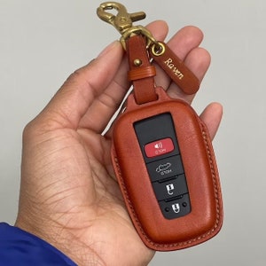 Può includere: Un portachiavi in ​​pelle marrone con una chiusura in metallo e un piccolo cartellino in pelle con la scritta "Raven". Il portachiavi ha una chiave nera con quattro pulsanti, tra cui un pulsante rosso con la scritta "Hold".