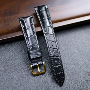 May include: Two black leather watch straps with a crocodile pattern. The straps have gold buckles and are laid out on a dark brown wooden surface.
