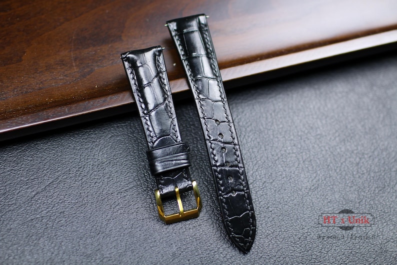 May include: Two black leather watch straps with a crocodile pattern. The straps have gold buckles and are laid out on a dark brown wooden surface.