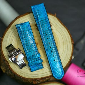May include: Two bright blue watch straps with a reptile skin pattern are displayed on a wooden slice. A silver watch clasp is positioned near the straps. The straps are a vibrant turquoise color, with visible stitching along the edges.