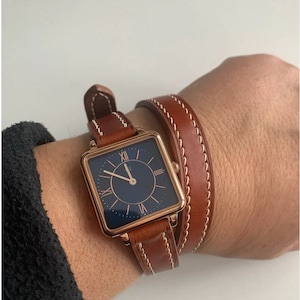 Puede incluir: Un reloj de forma cuadrada con una correa de cuero marrón y una esfera azul. El reloj tiene números romanos y una caja de color dorado.