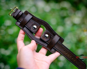 May include: A brown leather watch strap with a unique design featuring a cut-out section and two metal buckles. The strap is stitched with white thread and has a gold buckle.