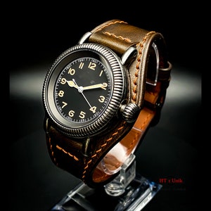 May include: A black-faced wristwatch with a brown leather strap. The watch has a silver case and a white dial with black numerals. The watch is on a display stand.