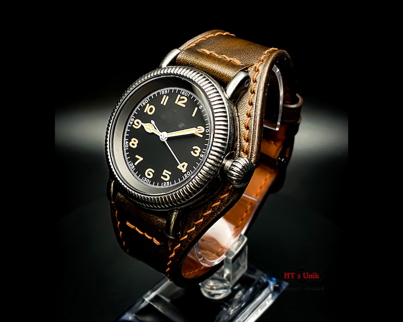 May include: A black-faced wristwatch with a brown leather strap. The watch has a silver case and a white dial with black numerals. The watch is on a display stand.