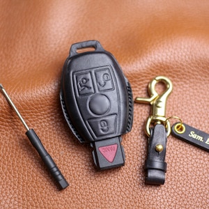 May include: Black leather car key fob cover with a key fob and a keychain with a gold-colored clasp. The keychain has a small black tag with the name "Sam. B" on it. A small screwdriver is also present.