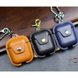 Puede incluir: Tres estuches de cuero para Airpods con llaveros. Los estuches son marrones, negros y azules. Los estuches tienen un pequeño bolsillo en la parte delantera. El texto "HT x Gnik" es visible en la esquina inferior derecha de la imagen.