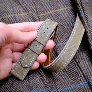 May include: A light grey leather watch strap with white stitching and a silver buckle. The strap is laid out on a brown and white patterned fabric.