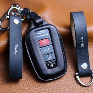May include: Black leather key fob with a silver key ring and a black leather strap with the name "Tracey" embossed on it. The key fob has a black leather cover with a silver button and a silver key ring. The key fob is on a brown leather surface.