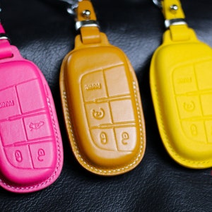Puede incluir: Tres fundas de cuero para llaves de coche. Las fundas son de color rosa, amarillo y marrón. Cada funda tiene un borde cosido y un diseño de llavero.