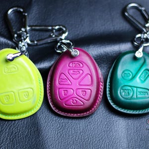 Peut inclure: Trois housses de clés en cuir de différentes couleurs : vert lime, magenta et sarcelle. Chaque housse a une disposition de boutons différente et est attachée à un porte-clés argenté.