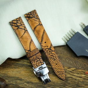 Puede incluir: Dos correas de reloj de cuero marrón con una textura similar a la de un reptil y marcas negras. Las correas están junto a herramientas de marroquinería, incluyendo un punzón de costura y una aguja de cuero. Las correas tienen un cierre plateado.