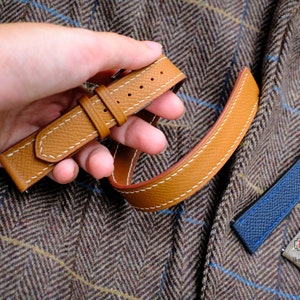 May include: A tan leather watch strap with white contrast stitching and a textured surface. The strap features two loops and a metal buckle. Additional watch straps in blue and gray are visible nearby, along with a logo reading HT x Unik.