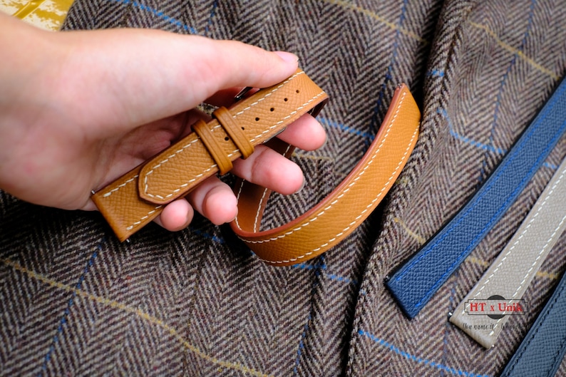 May include: A tan leather watch strap with white contrast stitching and a textured surface. The strap features two loops and a metal buckle. Additional watch straps in blue and gray are visible nearby, along with a logo reading HT x Unik.