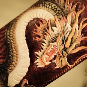 Op de afbeelding: Een bruine leren portemonnee met een gedetailleerd drakenontwerp. De draak is gesneden en beschilderd met tinten oranje, geel en wit, met rode accenten. De portemonnee heeft een textuur en een gestikte rand. De afbeelding bevat de tekst "HT x Unite".