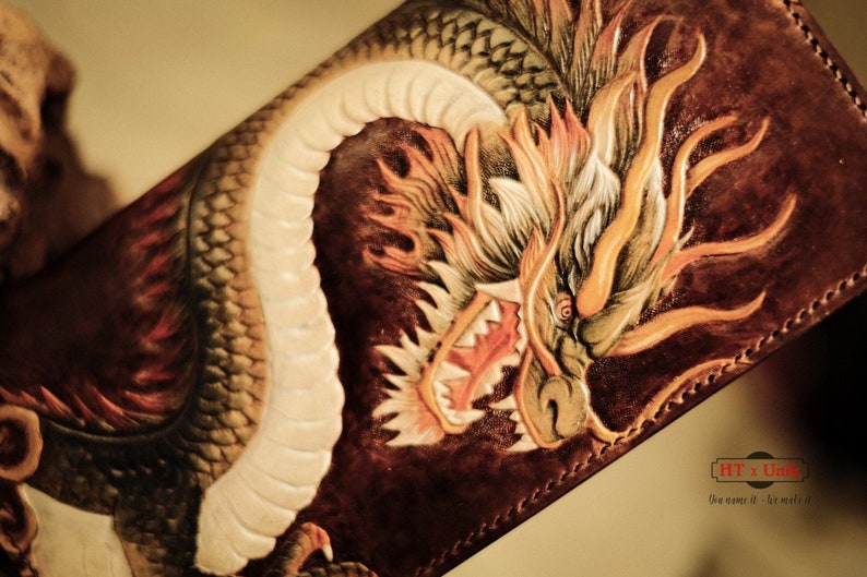 Op de afbeelding: Een bruine leren portemonnee met een gedetailleerd drakenontwerp. De draak is gesneden en beschilderd met tinten oranje, geel en wit, met rode accenten. De portemonnee heeft een textuur en een gestikte rand. De afbeelding bevat de tekst "HT x Unite".