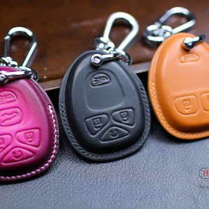May include: Three leather key covers in different colors: pink, black, and brown. Each cover has a silver metal ring and a silver metal hook. The covers are designed to fit over car key fobs.