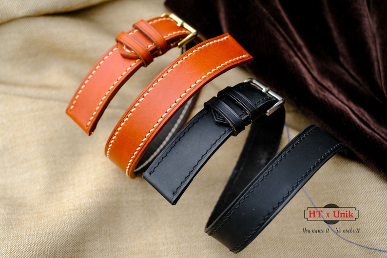 May include: Two leather watch straps, one in tan and one in black, each featuring contrast stitching and a metal buckle. The tan strap has a gold-tone buckle, while the black strap has a silver-tone buckle. A logo reads HT x Unik You name it We make it.