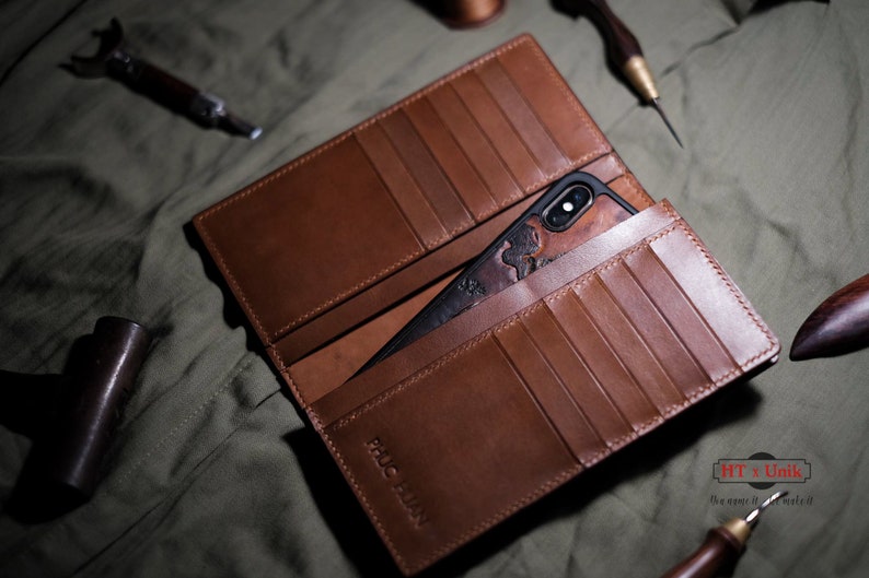 Op de afbeelding: Open bruin leren portemonnee met meerdere kaartsleuven en een telefoonhoesje. De telefoonhoes is zwart met een gesneden ontwerp. De portemonnee ligt op een groene ondergrond. De tekst "PHUC HUAN" is zichtbaar.