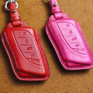 Puede incluir: Dos fundas de cuero para llaves, una roja y otra rosa, con un llavero plateado. Las fundas tienen un diseño de cuatro botones en la parte delantera.