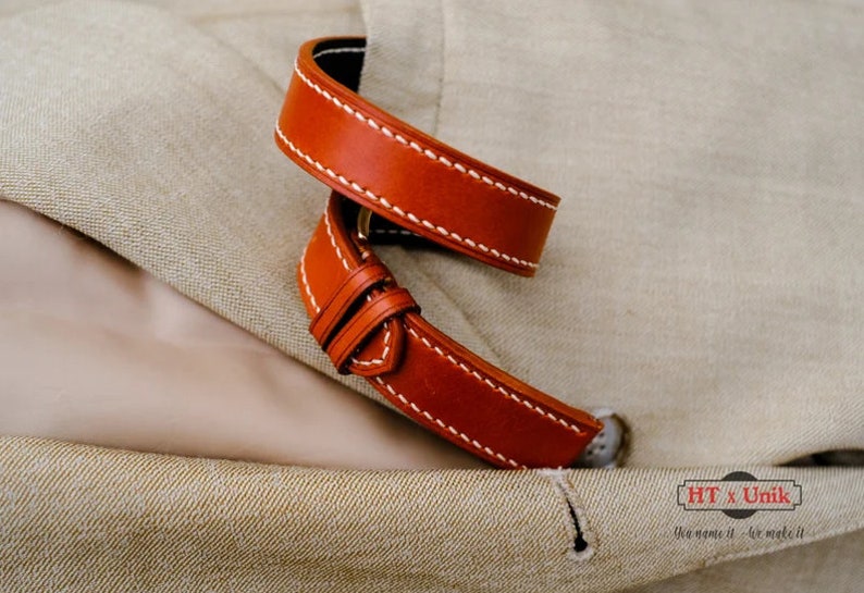 May include: A cognac-colored leather watch strap featuring white contrast stitching along the edges, a metal buckle, and two leather keepers. The strap is coiled and displays the text HT x Unik and the phrase You name it we make it.