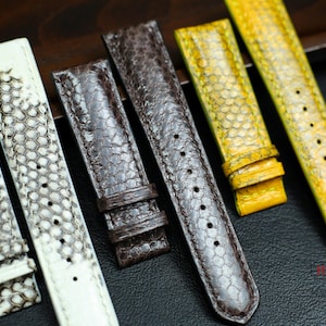 Könnte beinhalten: Eine Sammlung von sieben Uhrenarmbändern aus verschiedenen Materialien, darunter Leder, Schlangenhaut und ein gelber Stoff mit Muster. Die Armbänder sind in einer Reihe auf einer Holzoberfläche angeordnet.