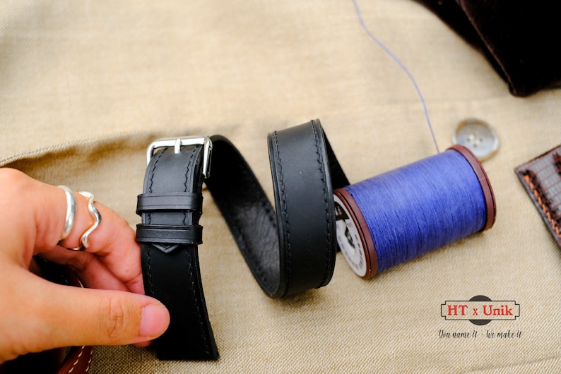 May include: A black leather watch strap with a silver-toned metal buckle and two keepers, positioned next to a spool of vibrant blue thread. The strap features visible stitching along the edges and a smooth finish, presented with a spool of thread.