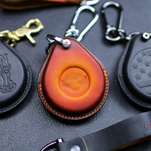 Könnte beinhalten: Drei Leder-Schlüsselanhänger mit verschiedenen Designs. Ein Schlüsselanhänger ist schwarz mit den Worten "Born to Ride" geprägt. Ein weiterer Schlüsselanhänger ist orange mit einem kreisförmigen Ausschnitt. Der dritte Schlüsselanhänger ist schwarz mit einem weißen und blauen amerikanischen Flaggen-Design.