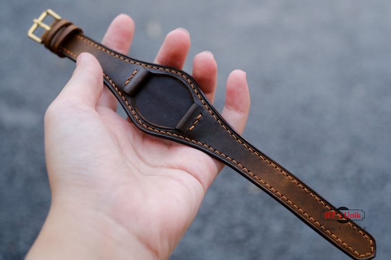 May include: A brown leather watch strap with orange stitching. The strap is shown from the back side and has a buckle on the left side.