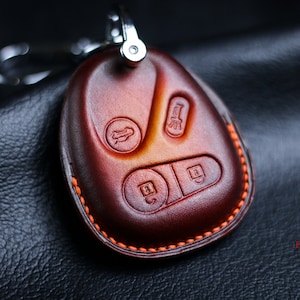 Puede incluir: Un llavero de cuero marrón con costuras naranjas. El llavero tiene cuatro botones con símbolos en relieve.