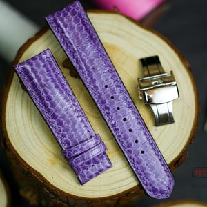 Może przedstawiać: Dwa paski do zegarka ze skóry w kolorze fioletowym z srebrną klamrą. Paski mają fakturowany wzór.