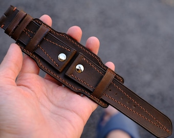 May include: A brown leather watch strap with orange stitching and silver metal accents. The strap is designed to be worn as a cuff bracelet.