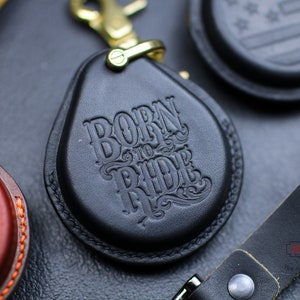 Puede incluir: Llavero de cuero negro con cierre de metal dorado y las palabras "Born to Ride" grabadas en relieve en la parte delantera.