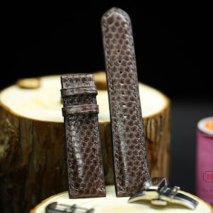 May include: Two brown leather watch straps with a textured pattern. The straps are on a wooden surface. The strap on the left is shorter than the strap on the right.