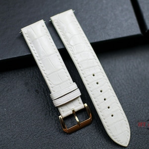 May include: Two white leather watch straps with a crocodile pattern. The straps have a gold buckle and are stitched with white thread.