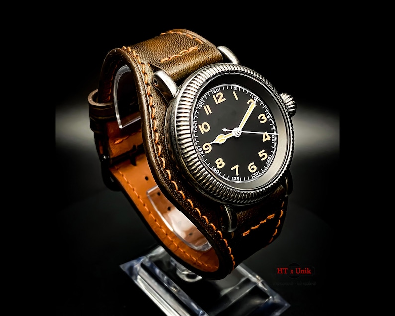May include: A brown leather watch strap with orange stitching wraps around a silver wristwatch with a black face and gold numerals. The watch face has a white second hand and a white minute hand. The watch face also has a white outer ring with black markings.