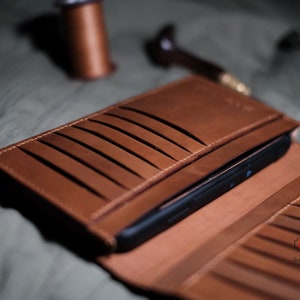 Op de afbeelding: Open bruin leren portemonnee met meerdere kaartsleuven en een zwarte smartphone erin. De portemonnee is open en toont het interieurontwerp. De achtergrond bevat leerbewerkingsgereedschap en -materialen.