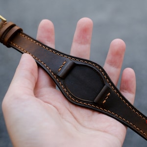May include: A brown leather watch strap with orange stitching. The strap is designed to fit a watch with a round face.