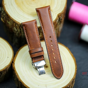 Può includere: Due cinturini per orologi in pelle marrone con cuciture arancioni e fibbia argentata sono esposti su una superficie di legno. I cinturini sono nuovi e inutilizzati. Sullo sfondo ci sono rocchetti di filo rosa e bianco.