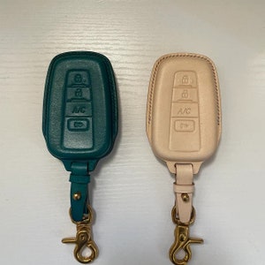 Può includere: Due copri chiavi in ​​pelle, uno verde acqua e uno beige chiaro, con portachiavi dorati. Entrambe le cover hanno pulsanti con la scritta "A/C" e altri simboli.