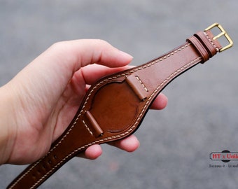 May include: Brown leather watch strap with white stitching and a gold-colored buckle. The strap has a unique design with a central pad and is held by a hand. The strap is a watch accessory.