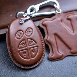 May include: Brown leather key chain with a silver key ring and a small, clear crystal. The keychain has a leather fob with four buttons and a separate leather charm with the letter "N" embossed on it. The text "HT x Unik" and "the name it - we make it" is printed on the charm.