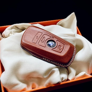 May include: A brown leather key fob with a silver button and four buttons. The key fob is in a white cloth box with an orange border.