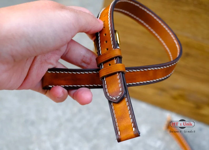 May include: A tan leather watch strap featuring dark brown edges and white contrast stitching. The strap includes a metal buckle and two leather keepers. A small logo in the corner displays the text HT x Unik and You name it - We make it.