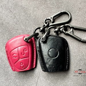 Könnte beinhalten: Zwei Leder-Schlüsselanhänger, einer in leuchtendem Pink und der andere in Schwarz, befestigt an Karabiner-Schlüsselanhängern aus Gunmetal. Die rosa Abdeckung hat geprägte Schlüsselsymbole. Die schwarze Abdeckung hat ein kreisförmiges Design.