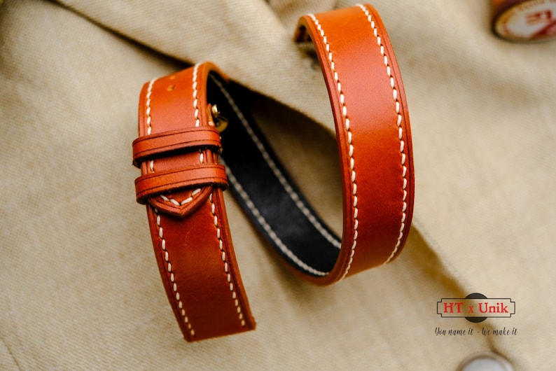 May include: A tan leather watch strap featuring white contrast stitching, two leather keepers, and a black interior lining. The strap is curved into a loop. A logo in the bottom right corner displays the text HT x Unik You name it - we make it.