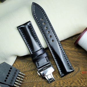 Könnte beinhalten: Zwei schwarze Lederarmbänder mit Steppnähten. Ein Armband hat eine silberne Schließe. Die Armbänder liegen auf einer Holzoberfläche.