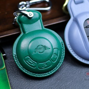 Può includere: Un portachiavi in pelle verde con un anello e un bottone argentati. Il portachiavi ha un design circolare con tre pulsanti e un logo al centro. Il logo è un cerchio stilizzato con una linea che lo attraversa.