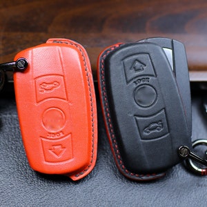 Op de afbeelding: Twee leren sleutelhangers met sleutelringen. Eén is rood en de andere is zwart. Beide hebben een auto-icoon en het woord "lock" erop.